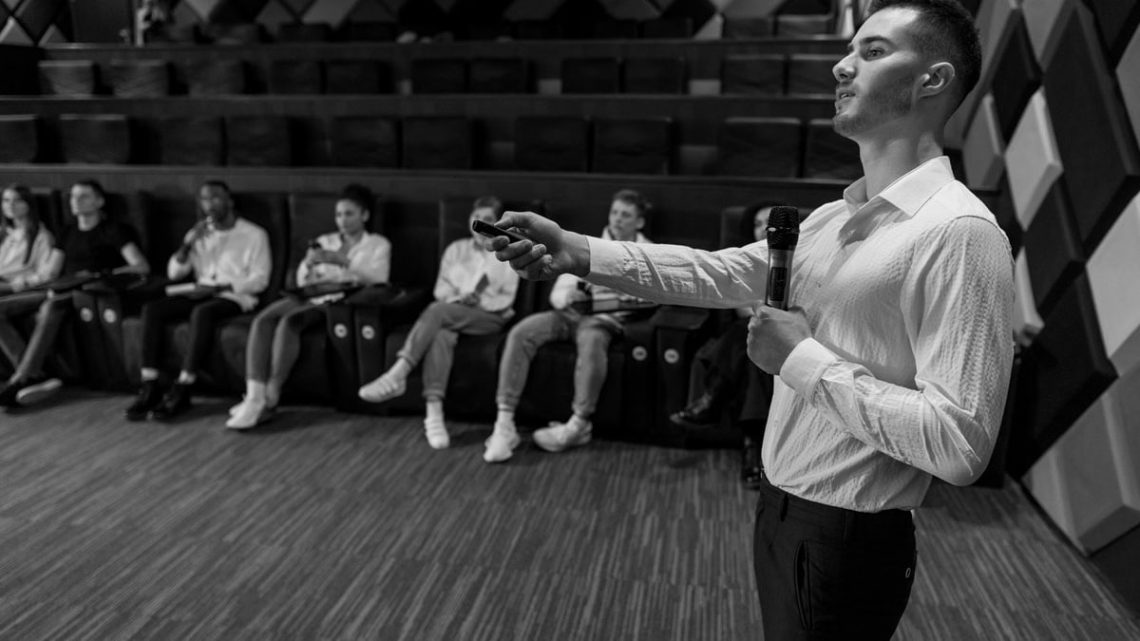 Monochromatic image of a speaker presenting to a small audience in a stylish auditorium.